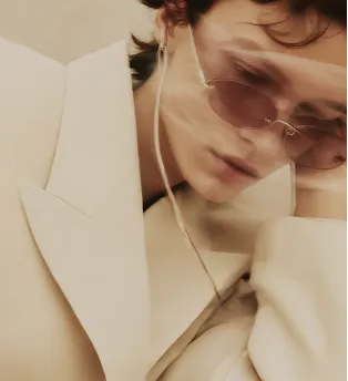 Person wearing beige coat and tinted sunglasses, resting their face on their hand.