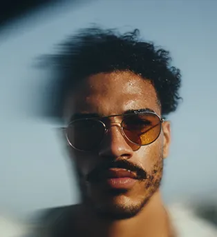 Portrait of a man with curly hair, mustache, and tinted sunglasses with blurred edges.
